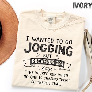 May include: An ivory short-sleeve t-shirt with black text that reads: I WANTED TO GO JOGGING BUT PROVERBS 28:1 SAYS THE WICKED RUN WHEN NO ONE IS CHASING THEM. SO THERE'S THAT. The shirt features a Comfort Colors brand label at the inner neckline.