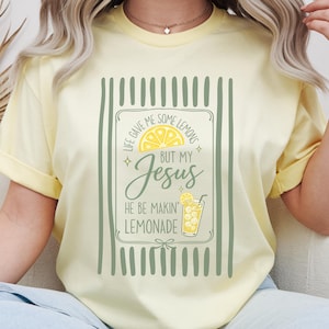 Può includere: T-shirt giallo chiaro con una grafica che dice "Life gave me some lemons but my Jesus he be makin' lemonade." La grafica include una fetta di limone, stelle e un bicchiere di limonata. La maglietta ha maniche corte.