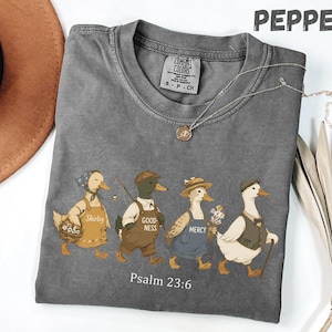 Könnte beinhalten: Ein graues T-Shirt mit Rundhalsausschnitt. Die Grafik zeigt vier Enten in Outfits, jede mit einem Wort auf ihrer Schürze. Der Text "Psalm 23:6" steht unter den Enten. Eine silberne Halskette liegt auf dem Shirt.