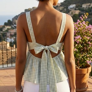Op de afbeelding: Een lichtgroene en witte geruite top met een strik op de rug. De top heeft brede bandjes en een peplum zoom. Het model draagt een witte broek. De achtergrond toont een stad en een plant in een pot.