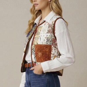Puede incluir: Un chaleco patchwork con ribete de cuero marrón y un bolsillo marrón. El chaleco presenta una variedad de parches de tela estampados florales en tonos crema, beige y colores apagados. La modelo lleva una camisa blanca con cuello y vaqueros azules.