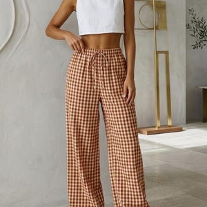Op de afbeelding: Een witte mouwloze crop top gecombineerd met een broek met hoge taille en wijde pijpen in een bruin en crèmekleurig ruitpatroon. De broek heeft een taille met trekkoord. De outfit wordt gecompleteerd met bruine sandalen.