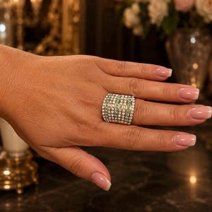 May include: A large, rectangular ring with a silver band and a surface covered in small, clear and green gemstones. The ring is worn on a finger, with a dark background and a blurred floral arrangement.