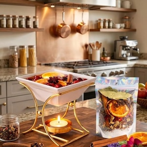 May include: A white square ceramic bowl sits on a gold metal stand with a lit tea light candle underneath. The bowl contains a liquid mixture with orange slices, cranberries, and cinnamon sticks. Next to it is a pouch of dried potpourri ingredients.