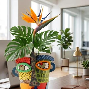 Peut inclure: Un pot de fleurs en céramique en forme de visage humain stylisé avec des motifs géométriques vifs rouges, jaunes, verts et bleus. Le pot contient une plante monstera verte et une fleur oiseau de paradis orange et bleue, créant un ensemble coloré.