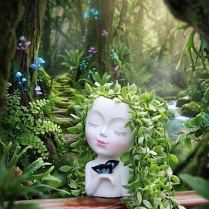 May include: A white head-shaped planter featuring closed eyes and pink lips, with a small blue butterfly resting on the hand. The planter is filled with trailing green foliage that cascades down the sides, creating a hair-like appearance.