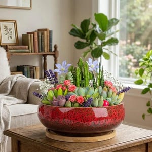May include: A round, deep red ceramic bowl planter with a textured, mottled finish, resting on a circular wooden base. The planter is filled with a variety of green succulents, small pink roses, purple lavender sprigs, and light blue flowers.