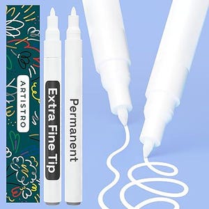 May include: Two white permanent paint markers with extra fine tips. The markers feature black labels with the text "Extra Fine Tip" and "Permanent". A dark green box with the brand name "ARTISTRO" and colorful scribbles is visible.