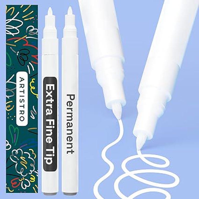 May include: Two white permanent paint markers with extra fine tips. The markers feature black labels with the text "Extra Fine Tip" and "Permanent". A dark green box with the brand name "ARTISTRO" and colorful scribbles is visible.