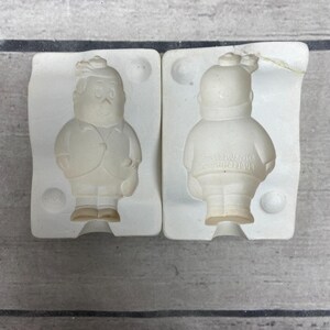 May include: Two off-white ceramic slip casting molds for a figurine of a person wearing a jacket and cap. The left mold shows the front of the figure, while the right mold shows the back with the embossed text "© 1984 Duncan" and "1984".