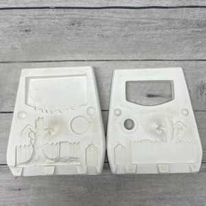 May include: Two off-white ceramic molds featuring a relief design of a graveyard scene. Each mold displays a gnarled tree, a picket fence, and a tombstone. The left mold is solid, while the right mold features a rectangular cutout at the top.