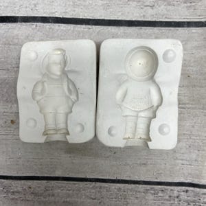 May include: Two white plaster molds, each depicting a child figure. One mold shows a boy in overalls and a jacket, while the other features a child in a hooded coat. The molds are set against a weathered, light-colored wooden surface.