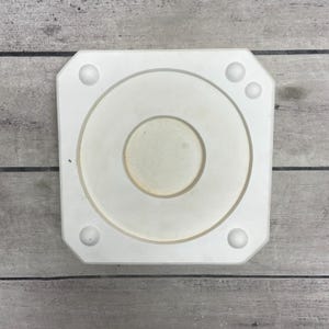 May include: A square white plaster mold featuring a central circular depression and a smaller inner circular indentation. The four corners of the mold are beveled and each corner contains a small, raised circular bump for alignment purposes.