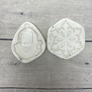 May include: Two off-white silicone molds for crafting. One mold is shaped like a teardrop and features a detailed embossed design of a Santa Claus face with a beard and hat. The second mold is hexagonal and features an embossed snowflake design.