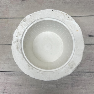 May include: A white circular plaster mold featuring a smooth interior basin with a grid pattern of small indentations. The exterior rim has a rough, uneven, and textured surface, contrasting with the clean, uniform design of the inner bowl.