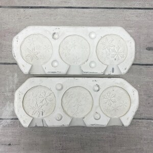 May include: Two rectangular white silicone molds, each featuring three circular cavities. Each cavity is embossed with intricate snowflake patterns of varying sizes. The molds have a smooth, matte finish with small circular indentations between the circles.