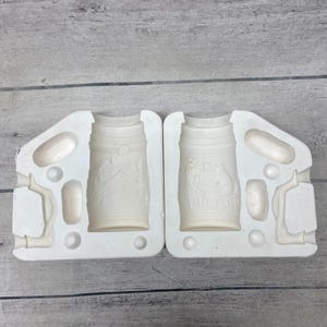 May include: A two-part white ceramic slip casting mold for a beer stein. The interior surfaces feature recessed relief designs of a house in a landscape and a group of people sitting at a table, along with separate cavities for handles.