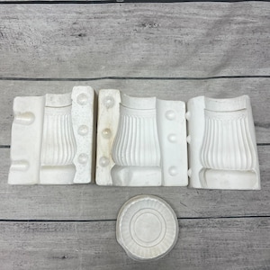 May include: Three white plaster casting molds featuring a vertical fluted design and a separate circular base mold. The molds have integrated registration pins and sockets for alignment, showing a concave interior shape for creating a fluted object.
