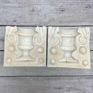 May include: Two off-white rectangular plaster molds for casting a decorative urn. Each mold features a central recessed urn shape with embossed leaf details, two side ring handles, and four circular indentations for alignment pins.