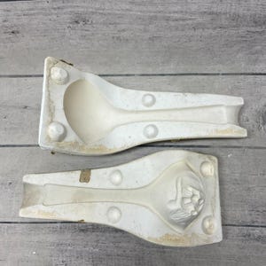 May include: Two off-white plaster mold halves for casting a spoon. The top piece features a concave spoon-shaped indentation, while the bottom piece displays a detailed relief carving of a floral motif at the bowl end, with circular registration points.