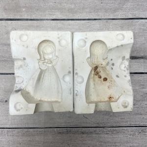 May include: Two white ceramic casting molds for a figurine of an angel with wings and a long dress. The left mold shows the front of the angel holding a small object, while the right mold shows the back of the angel with some brown discoloration.