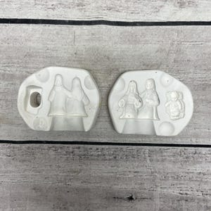 May include: Two white ceramic molds with religious figures. One mold features two figures, and the other has three figures, including an angel. The molds are on a light gray wooden surface.