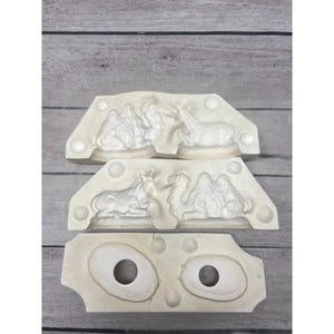 May include: Three off-white ceramic molds for casting figures. Two molds feature recessed relief designs of a camel and a resting animal. The third mold has two circular openings and circular indentations, designed for creating hollow shapes.