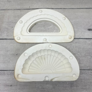 May include: Two off-white ceramic slip casting molds for pottery. The top mold features a semi-circular opening with a scalloped inner edge. The bottom mold displays a detailed fan-shaped relief pattern with radiating grooves and a central semi-circle.