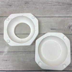 May include: Two white ceramic slip casting molds with an octagonal exterior shape. One mold features a large circular opening in the center, while the other is a solid bowl-shaped mold. Both pieces have four small circular indentations at the corners.