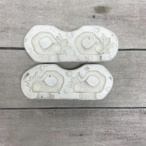 May include: Two off-white silicone or plastic molds, each featuring two recessed cornucopia designs. The cornucopia shapes are detailed with textured surfaces, including small pumpkins and corn cobs, arranged in a horizontal, symmetrical pattern.