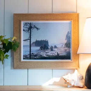 May include: A framed landscape print featuring a coastal scene with a rocky island, beach, and ocean. The frame has a woven texture. The print is displayed on a wooden surface, next to a lamp and a potted plant.