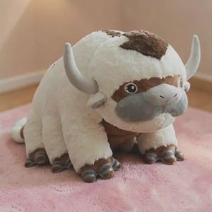 Puede incluir: Un peluche que representa un bisonte con cuernos grandes y curvados. El juguete es principalmente blanco con manchas marrones en la cabeza y los pies. Tiene un hocico gris y ojos grandes y expresivos. El juguete est&aacute; sobre una superficie rosa.