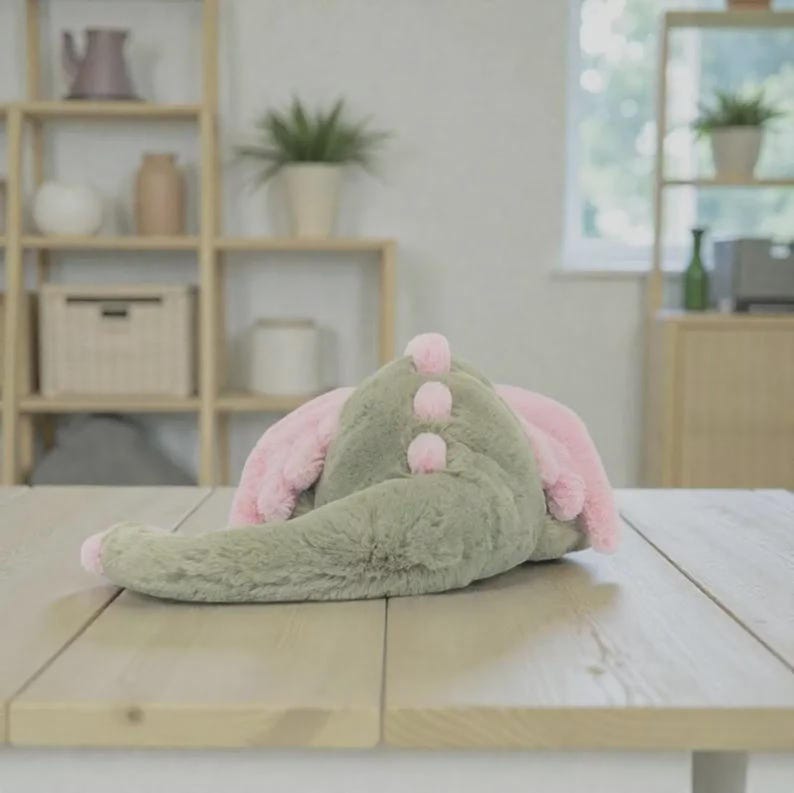 K&ouml;nnte beinhalten: Ein gr&uuml;nes Pl&uuml;sch-Dinosaurierspielzeug mit rosa Akzenten, einschlie&szlig;lich der Ohren und drei kleinen, runden Details auf dem R&uuml;cken. Das Spielzeug liegt auf einer hellen Holzoberfl&auml;che.