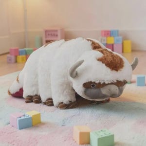 Puede incluir: Un peluche con forma de bisonte, con pelaje blanco y manchas marrones en la cabeza y la espalda. Tiene peque&ntilde;os cuernos grises y patas marrones. El juguete est&aacute; sobre una alfombra colorida, rodeado de bloques de juguete coloridos.