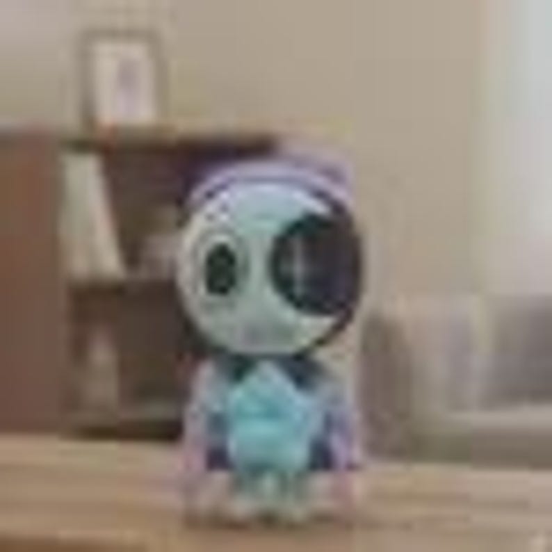 May include: A small, light blue and white figurine of a character with a large, round head and a cross-shaped design on its face. The figurine is wearing a blue and purple outfit and is sitting on a wooden surface.