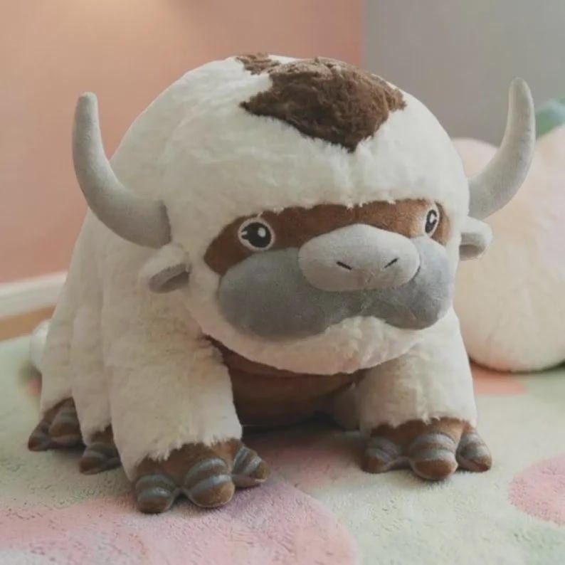 Puede incluir: Un peluche de una criatura blanca y esponjosa con una mancha marr&oacute;n en la cabeza, cuernos grises y un hocico gris. El juguete tiene ojos grandes y expresivos y pies detallados con detalles marrones y grises.