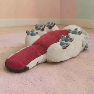 Puede incluir: Una zapatilla de felpa con forma de pie de criatura, con una parte central roja, laterales blancos y huellas de patas azules. Fabricada en material suave y afelpado, descansa sobre una superficie rosa.