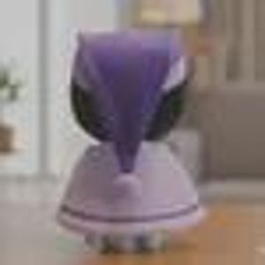 May include: A purple and white head massager with a rounded base and a top section that resembles a stylized owl. The massager has soft, rounded bristles and is designed for scalp massage. The background is blurred, suggesting an indoor setting.