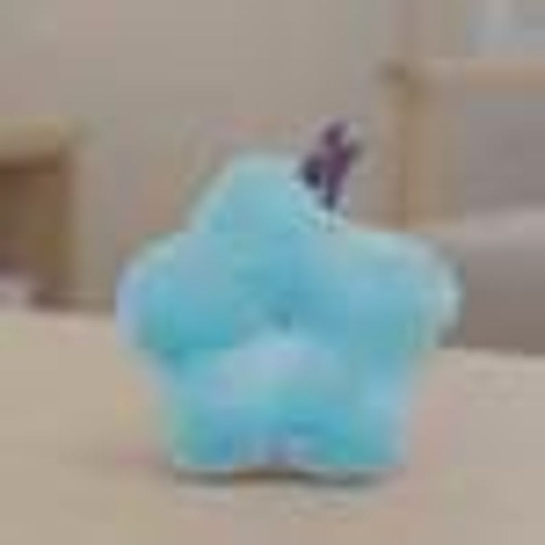 May include: A light blue, star-shaped plush toy with a small purple stem. The soft, fluffy toy is sitting on a light-colored surface. The toy is a whimsical decorative item.