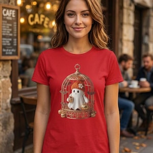 Könnte beinhalten: Ein rotes Kurzarm-T-Shirt mit einem Grafikdruck eines weißen Geistes, der eine orangefarbene Strickmütze mit einer weißen Blume trägt und eine Kaffeetasse in einem goldenen, verzierten Vogelkäfig mit kleinen Fledermäusen und Kürbissen hält.