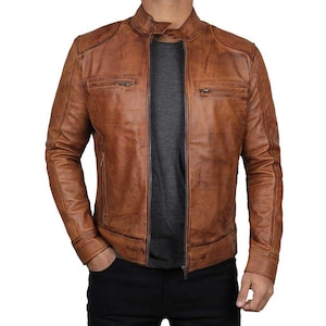May include: A tan leather jacket with a distressed finish, featuring a mandarin collar with a snap button closure, a full-length front zipper, and two horizontal zippered chest pockets. The jacket has long sleeves with cuffs and a fitted silhouette.