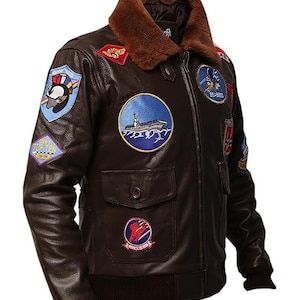 May include: Brown leather bomber jacket with a brown faux fur collar. The jacket features multiple embroidered patches, including a ship, eagle, and other military-themed designs. It has a ribbed hem and cuffs, and two front pockets.