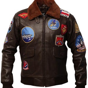 May include: Brown leather bomber jacket with a brown faux fur collar. The jacket features multiple embroidered patches, including "Texas Dallas", "Seabees", and "Tomcat". It has two front pockets and a ribbed hem and cuffs. The jacket is a classic aviator style.