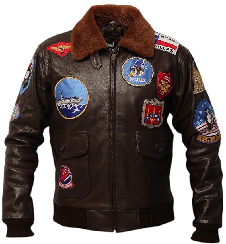 May include: Brown leather bomber jacket with a brown faux fur collar. The jacket features multiple embroidered patches, including "Texas Dallas", "Seabees", and "Tomcat". It has two front pockets and a ribbed hem and cuffs. The jacket is a classic aviator style.