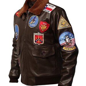 May include: A brown leather bomber jacket with a brown fur collar. The jacket features multiple embroidered patches, including "Texas Dallas", "Seabees", and "Tomcat". It has two front pockets and a ribbed cuff and hem.