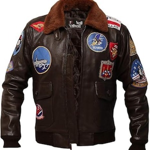 May include: A brown leather jacket with a brown fur collar and multiple patches. The jacket has a zipper closure, two front pockets, and ribbed cuffs and hem. Patches include military-themed designs and text.