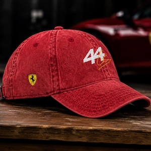 May include: Red baseball cap with the number "44" in white and a gold signature. The cap features the Ferrari logo on the side and an adjustable strap. The cap is made of a textured material.