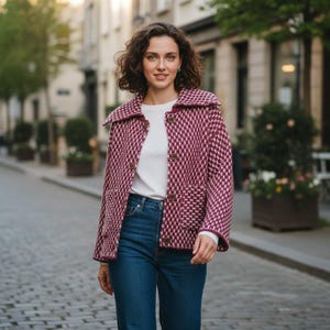 Peut inclure: Veste à carreaux bordeaux et blancs avec un large col et des boutons en bois. La veste est portée sur un haut blanc à manches longues et associée à un jean bleu. La personne marche dans une rue pavée.