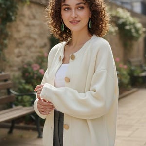 Peut inclure: Cardigan crème avec de gros boutons ronds en bois. Le pull à manches longues a un ourlet et des poignets côtelés. La personne porte un haut blanc et un pantalon gris. L'arrière-plan comprend un mur en pierre et de la verdure.