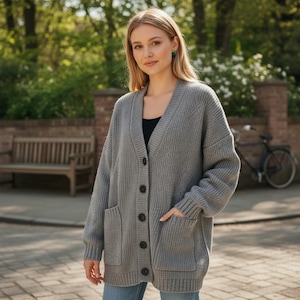 Peut inclure: Un cardigan gris en maille épaisse avec un col en V, une fermeture boutonnée et deux poches avant. Le cardigan est porté sur un haut noir et associé à un jean bleu. Le modèle se tient à l'extérieur sur une allée pavée de briques.